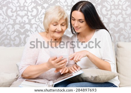 Old memories. Smiling granddaughter and her grandmother sitting on couch and ad looking at old pictures.