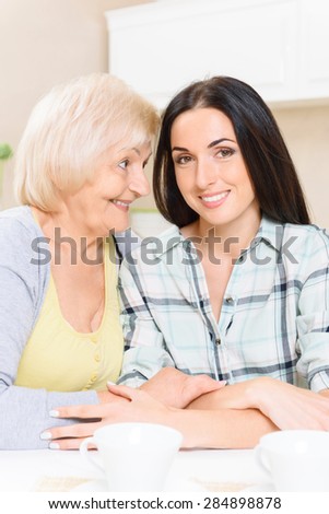Cute pair. Two young and old ladies sitting in kitchen embracing and talking.