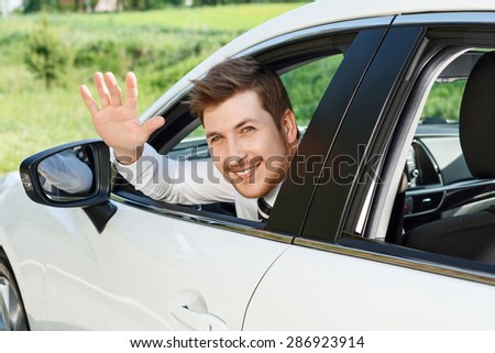 Saying hello. Handsome young smiling businessman sitting in new car and waving his to someone.