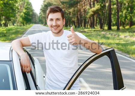 It is fine. Young handsome man standing near car with opened door and thumbing up.