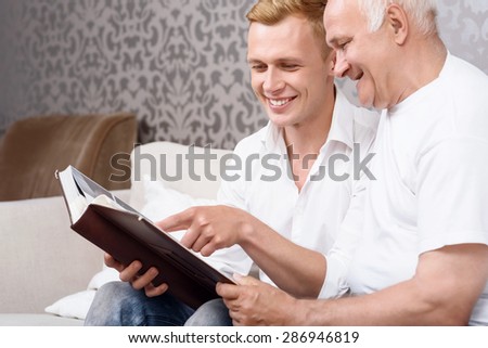 Look here. Young grandson sitting on couch together  with his grandfather and pointing at photo from album