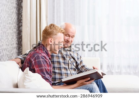 Old photos. Grandfather and grandson sitting on couch holding album and looking through old pictures