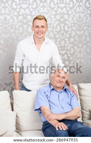Family photo. Young handsome grandson standing behind his old grandfather sitting on couch
