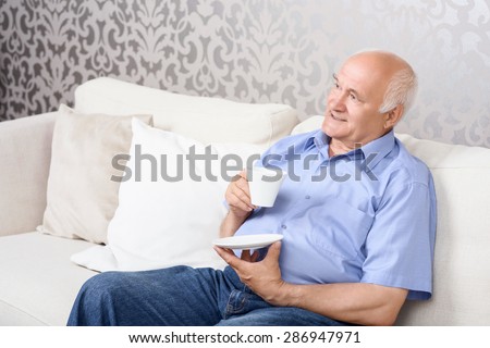 Coffee break. Thoughtful old man sitting on couch and holding white cup.