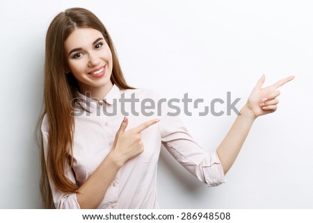 Portrait of a young beautiful girl with long brown hair wearing pink cotton blouse, standing waist up smiling and pointing with two hands to the left, on a white background