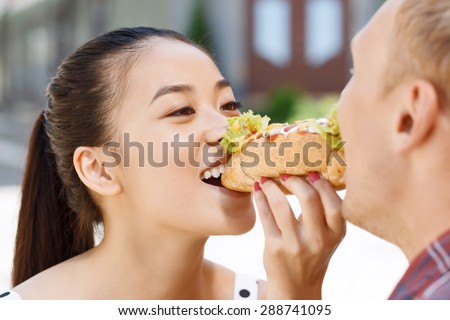 Eating together. Young handsome man and pretty woman standing and eating one hotdog.