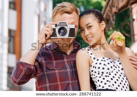 Old-schooled camera. Portrait of young man standing near his girlfriend holding hotdog and taking picture by using old camera.
