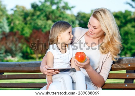 Learning with technologies. Pretty blond-haired mother sitting  on bench with her daughter holding tablet and apple