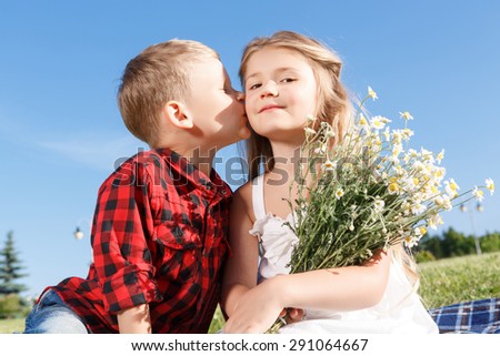 Puppy love. Positive little boy keeping his eyes closed and kissing the girl with flowers while sitting on blanket.