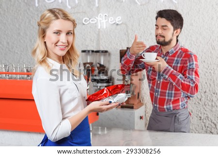 Feeling satisfaction. Portrait of pretty young blond-haired barista standing with coffee packages on background of satisfied customer.