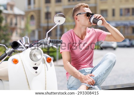 Pit stop. Young handsome blond-haired man sitting on bench near his scooter and drinking coffee.