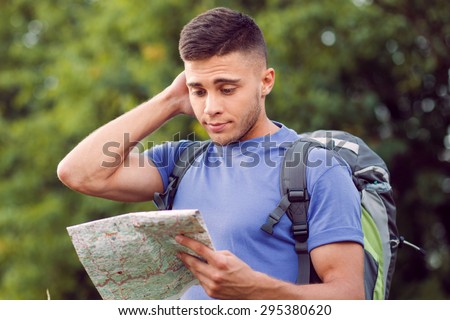 Traveling. Portrait of a young handsome tourist wearing blue t-short and backpack standing, feeling lost scratching head and looking at the map in his hand