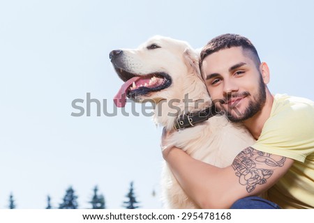 Portrait of a handsome man wearing yellow t-short and jeans with tattoo on his arm sitting on the grass, looking at us smiling and hugging his lovely golden retriever in the park