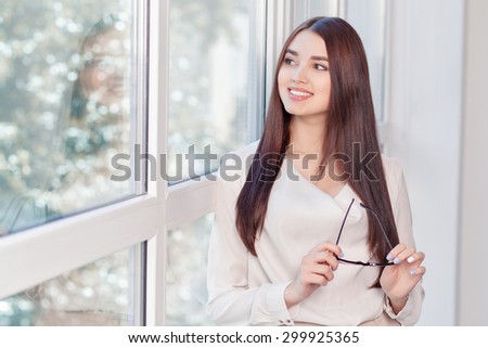 Let me think.  Smiling positive business woman holding glasses and looking aside while standing near window.