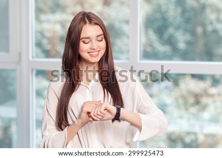 Do not hurry. Upbeat positive business woman keeping glance down and looking at her wrist watch while standing near window