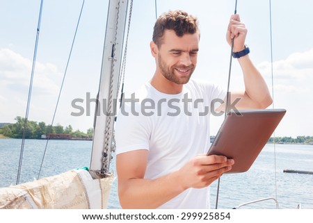 Time to check email. Pleasant smiling man keeping rope and holding laptop while surfing the Internet on board of yacht.