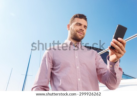 Waiting for good news. Positive smiling businessman holding mobile phone and going to make call while standing near his yacht