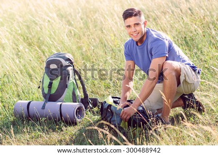 Green tourism.  Portrait of a young handsome tourist wearing blue t-short and beige shorts, sitting in the field fastening his bag smiling