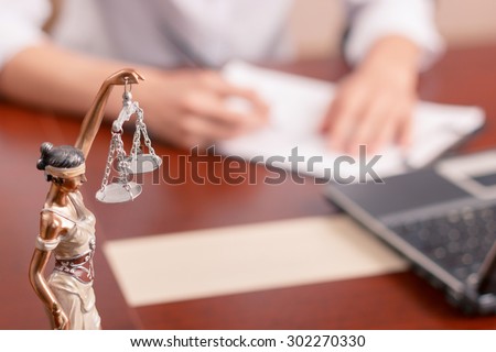 Follow the law. Professional lawyer sitting at the table and signing papers with justice statue standing on surface in forefront.