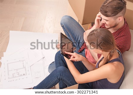 Prepare for living. Pleasant young wife sitting near settee and holding laptop while pointing it to her husband talking on mobile phone.