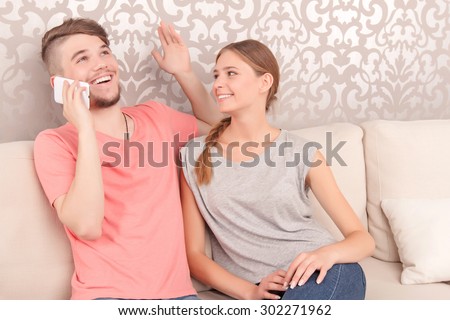 What a great news.  Jubilant smiling young husband sitting on the sofa with his wife and having conversation while holding mobile phone.