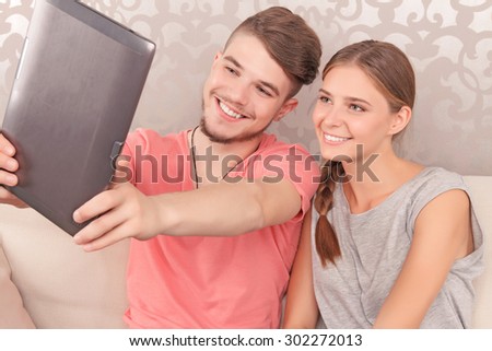Lets make photo. Smiling elated young couple holding laptop and making selfie while sitting on the sofa.