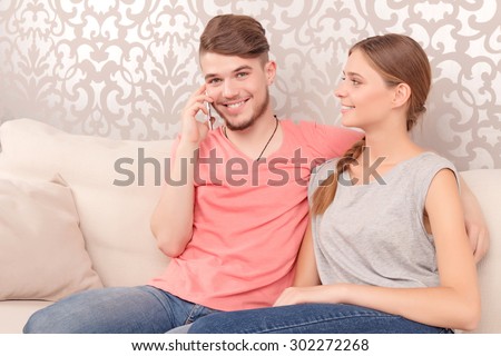 Brisk conversation. Pleasant elated  young husband sitting with his wife on the couch and holding mobile phone while talking
