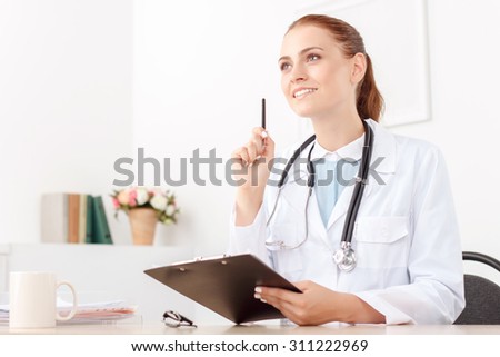 Clear mind. Pleasant lively pretty doctor woman holding folder with pen and sitting at the table while thinking out.