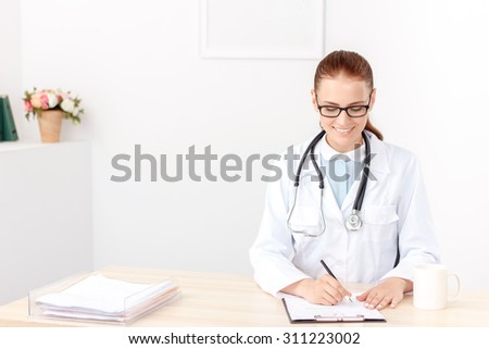Happy at work. Upbeat smiling doctor sitting at the table and writing while being busy at work