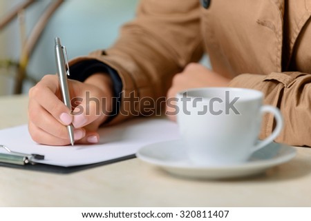 Get inspiration. Close up of cup of coffee standing on the table and nice young woman holding pen and writing