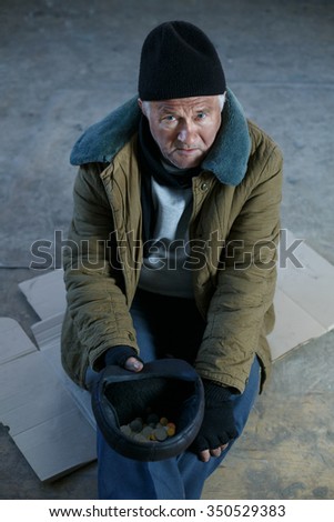Asking for help. Depressed old beggar is sitting on cardboard and holding his cap to ask for money.