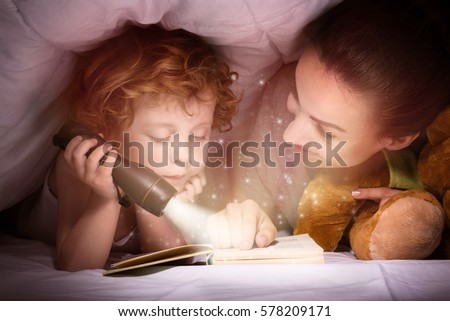 Time for stories. Delighted happy cute mother and son enjoying a book before sleeping and using a flashlight.