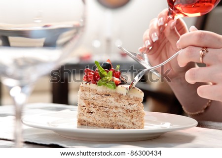 A piece of cake with forest berries decorated with mint leaf on a white plate in a restaurant, a woman sitting, holding a glass of wine
