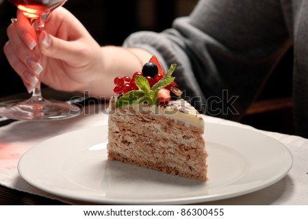A piece of cake with forest berries decorated with mint leaf on a white plate in a restaurant, a woman sitting, holding a glass of wine