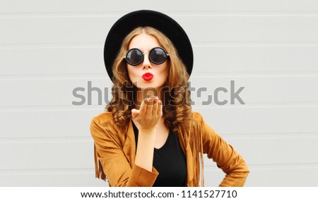 Pretty woman sends air sweet kiss wearing a black hat, sunglasses and brown jacket outdoors over urban grey background