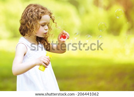 Cute curly little girl blowing soap bubbles outdoors in summer day