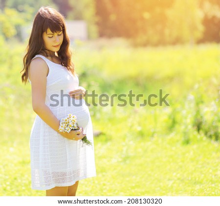 Pregnancy lovely woman in white dress with camomile flowers in summer sunny day, tender moment