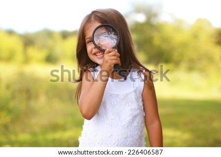 Positive cheerful little girl looking through a magnifying glass outdoors