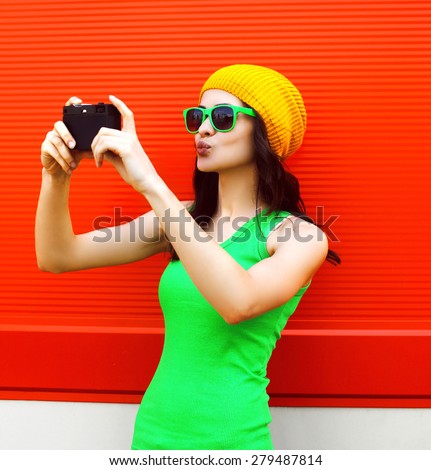 Fashion, summer, technology and people concept - pretty cool girl in sunglasses makes self-portrait on the camera outdoors against colorful red wall