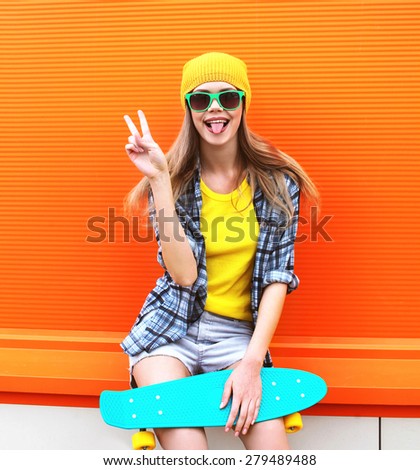 Fashion portrait of hipster cool girl in sunglasses and colorful clothes with skateboard having fun against the orange wall