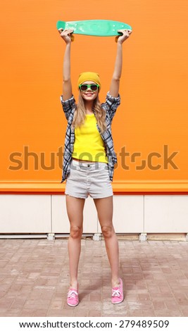 Fashion cool girl in sunglasses and colorful clothes with skateboard having fun against the orange wall
