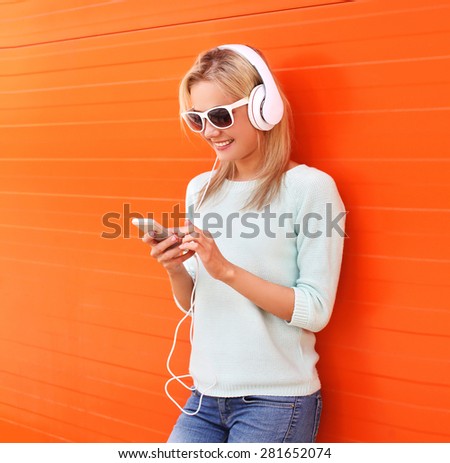 Fashion, technology and people concept - pretty smiling girl listen to music in headphones and using smartphone against the colorful orange wall