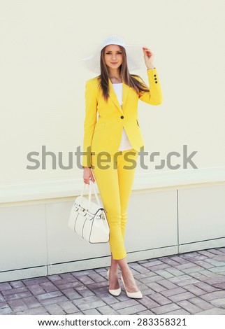 Beautiful young woman in yellow suit clothes and hat with handbag outdoors, street fashion