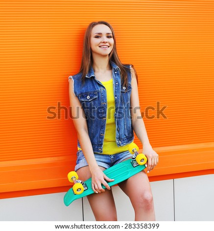 Fashion portrait of hipster girl in colorful clothes with skateboard having fun against the orange wall