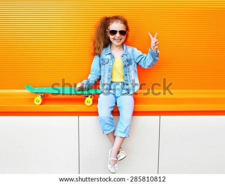 Fashion kid. Stylish little girl child wearing a jeans clothes and sunglasses with skateboard having fun in city and enjoys summer against the colorful orange wall