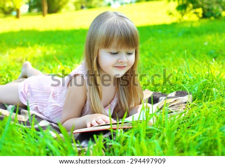 Portrait of little smiling girl child reading a book lying on the grass in sunny summer day