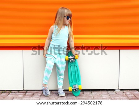 Fashion kid concept - stylish little girl child wearing a t-shirt and sunglasses with skateboard in the city summer against the colorful orange wall