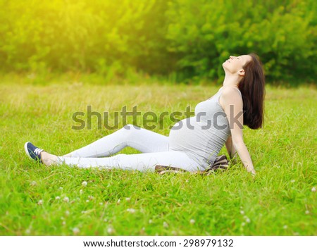 Happy pregnant woman sitting on the grass and enjoys sunny summer day