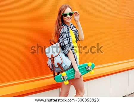 Fashion happy smiling hipster cool girl in sunglasses with skateboard and backpack having fun against the colorful orange wall
