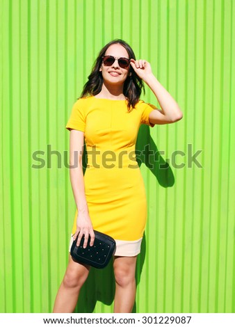 Beautiful young woman in yellow dress with black handbag clutch over green wall, street fashion